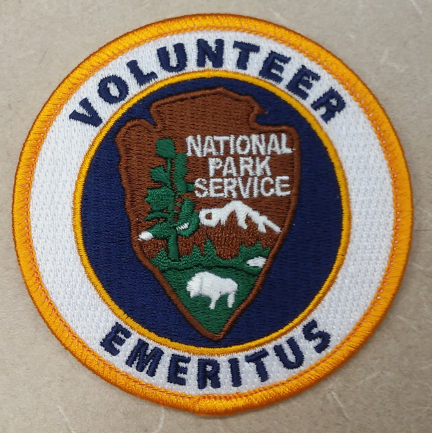 Runder Aufnäher mit blauem Rand und weißem Hintergrund, der mittig den schwarzen Text 'Volunteer National Park Service Emeritus' in einem gelben Kreis zeigt.
