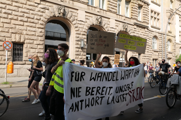 Eine Gruppe von Menschen marschiert auf einer Demonstration durch die Straße, einige halten Schilder und andere fahren Fahrröder, im Hintergrund ein Gebäude mit Fenstern, Überdachungen, Pfeilern und Skulpturen.