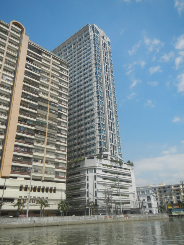 Ein hohes Wohngebäude am Wasser mit Bäumen, Pfählen und anderen Gebäuden im bewölkten Himmel.