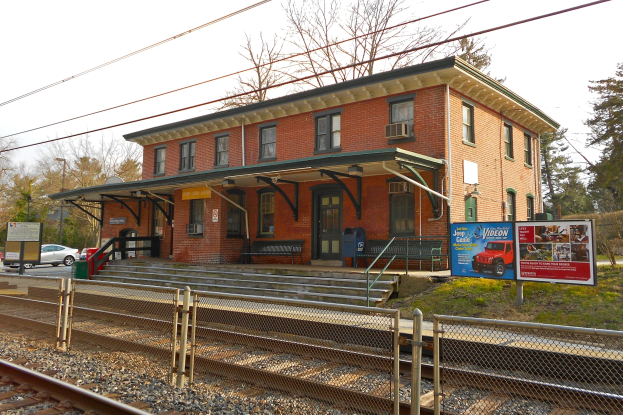 Ein Bahnhof mit einem Zug auf den Schienen, ein Gebäude mit Fenstern und Türen, Bänke, Informationsschilder, Maschendrahtzaun, Gras, Bäume, Fahrzeuge auf der Straße, Laternenmasten, Drähte und ein Himmel im Hintergrund.