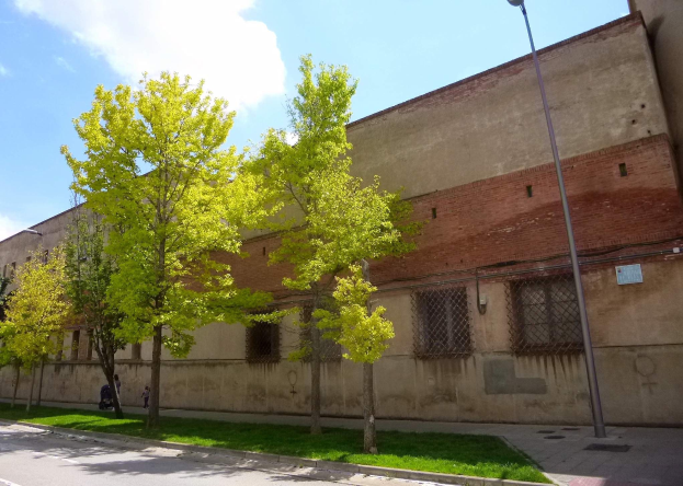 Altes Gebäude in Madrid, Spanien, mit einem Baum davor, umgeben von Straßenmasten, Laternen, Fahrzeugen, Passanten und Gittern, vor einem bewölkten Himmel, als ehemaliges Gefängnis zum Verkauf angeboten.