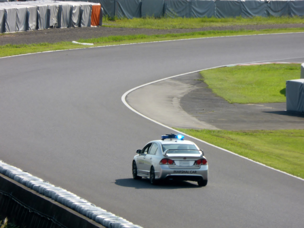 Ein Polizeiauto fährt eine Rennstrecke entlang, mit Gras auf beiden Seiten und Menschen, Zelten und anderen Gegenständen im Hintergrund.