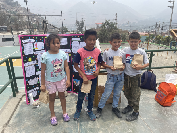 Kinder auf einem Basketballfeld mit Papiertüten in den Händen, mit Materialien auf dem Boden und einer Tafel mit Text links daneben, vor dem Hintergrund von Zäunen, Pfosten, Bäumen, Gebäuden, Hügeln und dem Himmel.