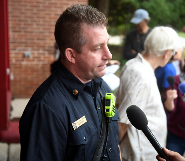 Ein Polizist in Uniform spricht mit Reportern, die Mikrofone halten, vor einer Backsteinwand und Bäumen.
