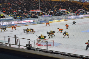 Eine Gruppe von Menschen, die Hockey auf einem Eisring in einer Arena spielen, mit einem Netz, einer Umzäunung, sitzenden Zuschauern, Bannern und Scheinwerfern von oben.
