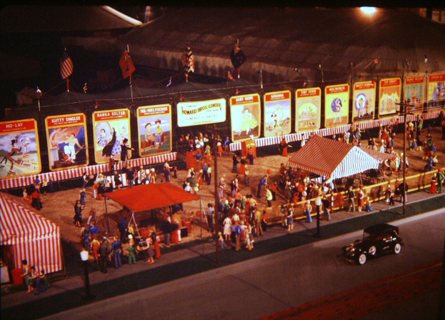 Ein altes Nachtfoto eines Faschings mit Menschen, einem Auto, Pfölen, einem Zaun, einem Zelt, Brettern mit Bildern und Text sowie Flaggen und einer Straßenlaterne.