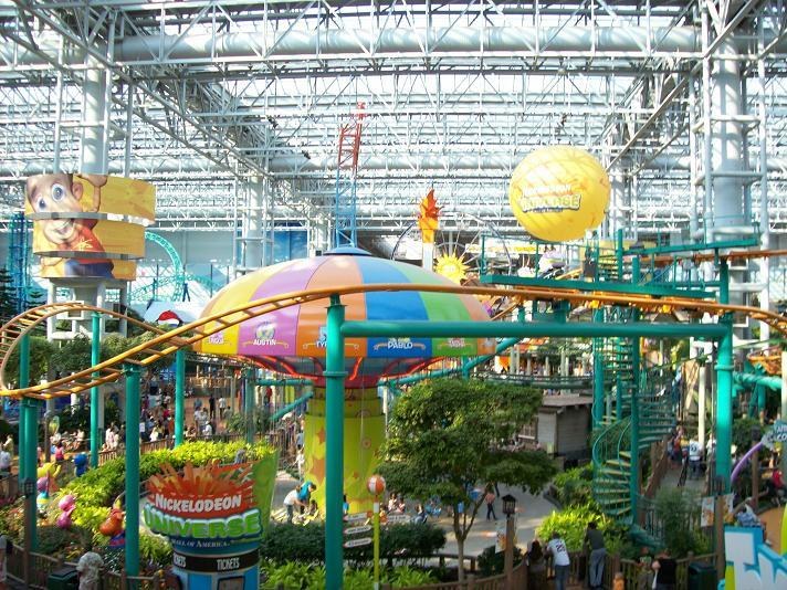 Ein großer Indoor-Wasserpark mit einer Achterbahn, umgeben von Menschen, Bäumen, Pflanzen und einer lebendigen Beleuchtung von oben.