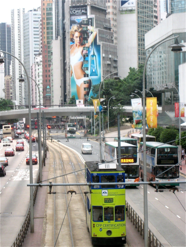 Eine belebte Stadtstraße mit verschiedenen Fahrzeugen, darunter Doppeldeckerbusse, Autos und andere Fahrzeuge, unter einem klaren blauen Himmel, mit hohen Gebäuden, einer Brücke und Beschilderung im Hintergrund.