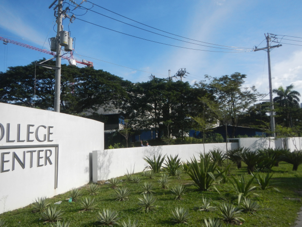 College Center-Eingangsschild an einer Gebäudewand, umgeben von Grün, einem Weg, Strommasten, Bäumen, fernen Gebäuden, einem Kran und einem bewölkten Himmel.