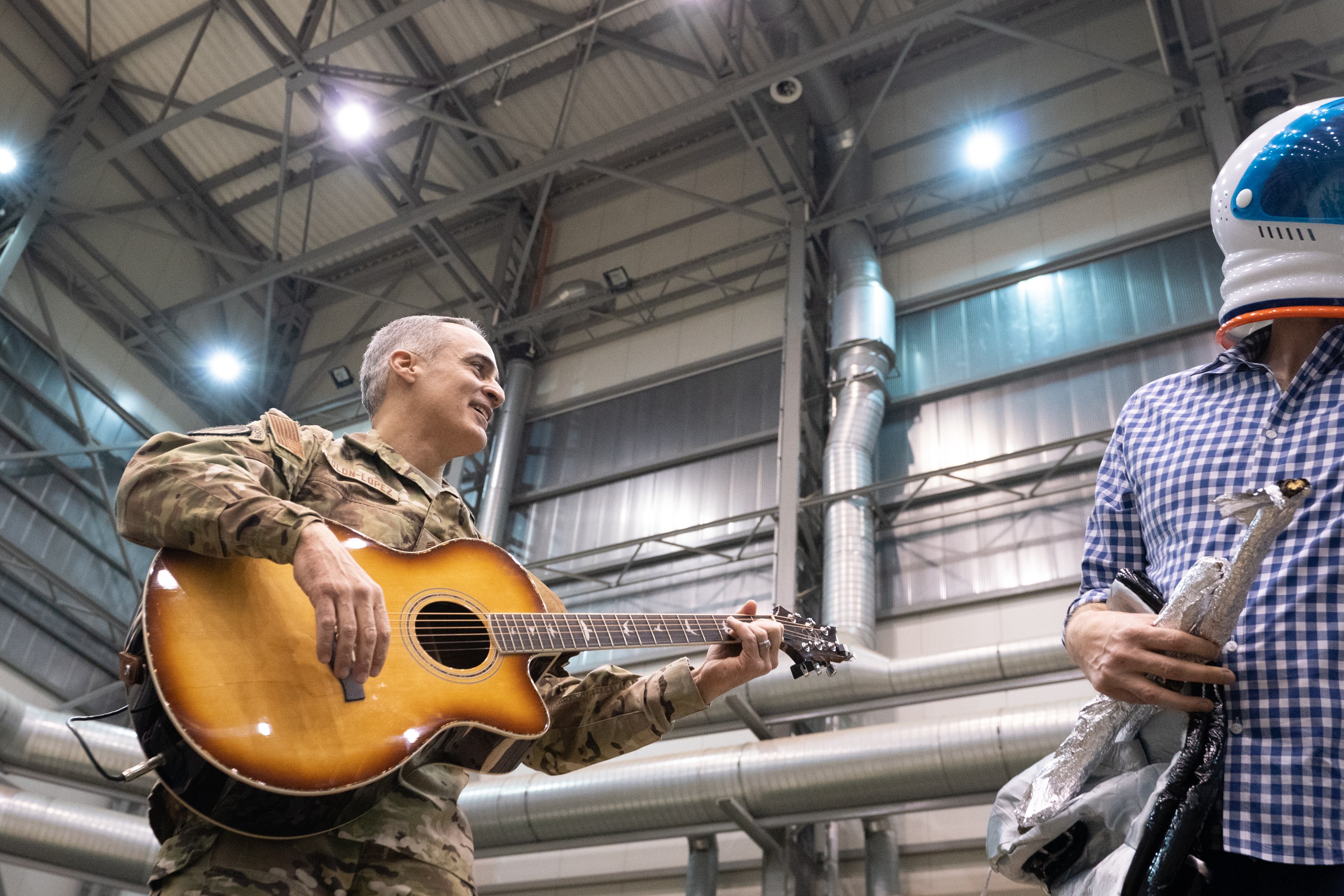 Ein Mann in Militärkleidung spielt eine Gitarre neben einem anderen Mann in einem Helm, mit Rohren und Lichtern im Hintergrund, was auf eine Militärband-Situation hinweist.