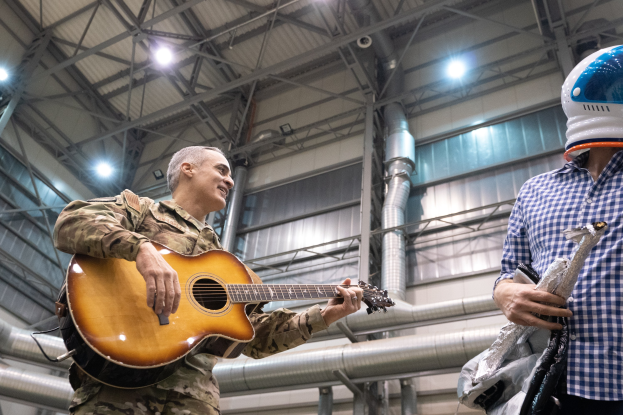 Ein Mann in Militärkleidung spielt eine Gitarre neben einem anderen Mann in einem Helm, mit Rohren und Lichtern im Hintergrund, was auf eine Militärband-Situation hinweist.