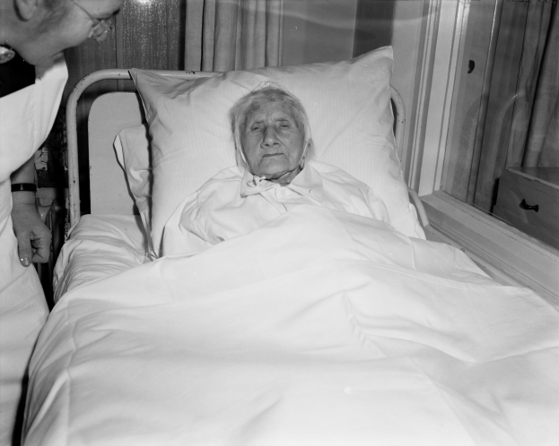 Eine ältere Frau liegt in einem Krankenhausbett mit einer weißen Bettdecke und Kissen, während eine Krankenschwester neben ihr steht, mit einem Fenster mit Vorhängen im Hintergrund.