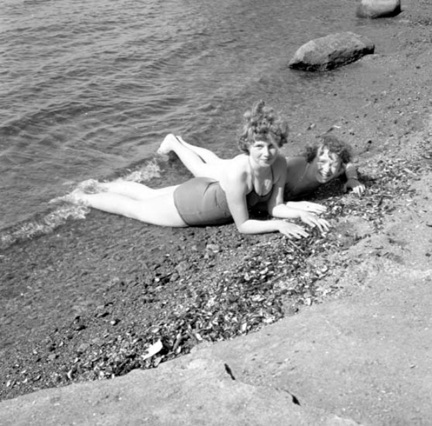 Zwei Frauen in Badeanzügen liegen auf einem Strand umgeben von Steinen und glitzerndem Wasser, dargestellt in Schwarz-Weiß.