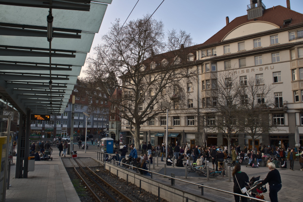 Eine belebte Stadtstraße mit einem Bahnhof auf der linken Seite, umgeben von Gebäuden, Bäumen, Laternenmasten und Texttafeln, wo Menschen gehen, auf Bänken sitzen oder stehen.