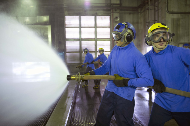 Mannergruppe in blauen Hemden und gelben Helmen bei der Arbeit an einer Maschine in einer Fabrik mit sichtbaren Rohren, Fenstern und Beleuchtung, einer Person sprüht Wasser auf den Boden.