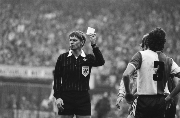 Schwarzes und weißes Bild eines Schiedsrichters mit roter Karte, mit einer Menge Menschen in Fußballtrikots dahinter einem Zaun.