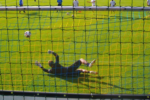 Ein Fussballer hält den Ball vor dem Tor, mit einem Netz im Vordergrund und einigen Menschen im Hintergrund sowie mehreren Bällen auf dem Boden verteilt.