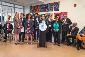 Eine Gruppe von Menschen steht und sitzt in einem Raum mit einem Podium, Mikrofon und einer Tafel mit der Aufschrift "Sancho Stadtverordnetenvorsteher tritt dem Stadtverordnetenkollegium bei", mit Flaggen, Feuerlöscher und Deckenleuchten an der Rückwand.