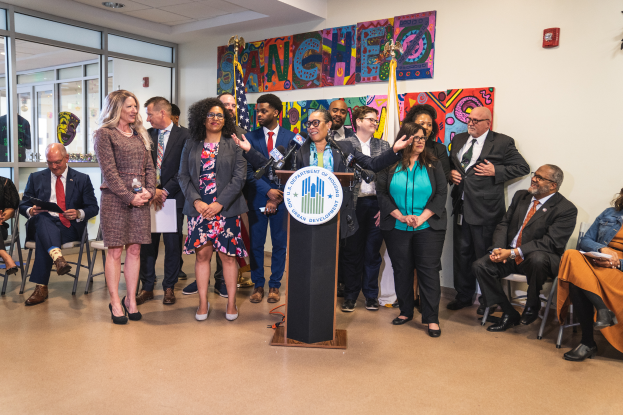 Eine Gruppe von Menschen steht und sitzt in einem Raum mit einem Podium, Mikrofon und einer Tafel mit der Aufschrift "Sancho Stadtverordnetenvorsteher tritt dem Stadtverordnetenkollegium bei", mit Flaggen, Feuerlöscher und Deckenleuchten an der Rückwand.