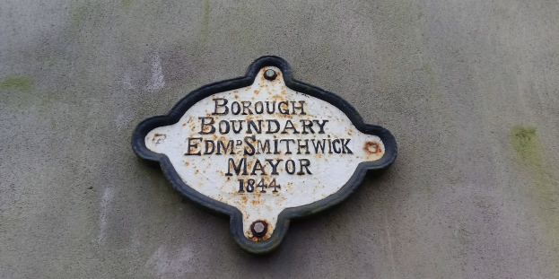 Metallplatte, die die Borough-Grenze markiert, mit 'Borough Boundary Edm Smithwick Mayor' in fetter weißer Schrift auf schwarzem Hintergrund.