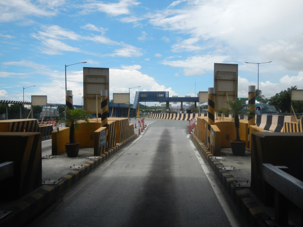 Straße mit einer Mäßstation, Absperrpfählen, Verkehrskegeln, Topfpflanzen, Hinweisschildern, Straßenmasten, Bäume und eine Brücke.