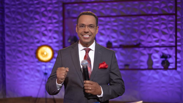 Stephen Colbert, Moderator von *The Daily Show with Trevor Noah*, steht auf einer Bühne und hält ein Mikrofon, trägt einen Anzug, Krawatte und Brille, mit einem Scheinwerfer und Ständer im Hintergrund.