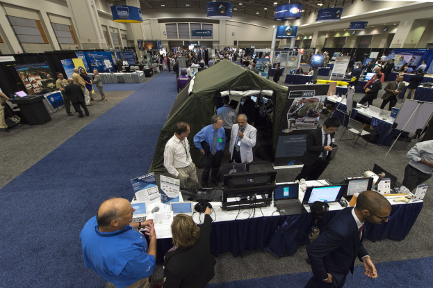 Große Gruppe von Menschen auf einer Karriere-Messe um einen Tisch mit Laptops, Monitoren und Papieren versammelt, mit einem Zelt, Schautafeln, Deckenleuchten und Fenstern im Hintergrund.
