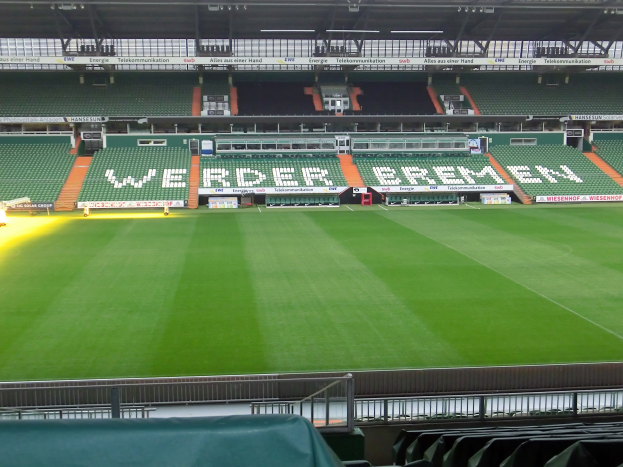 Großes Stadion mit einem grünen Feld, umgeben von Sitzplätzen, Werbetafeln, Beleuchtungsstrukturen und Geländern, mit einem drapierten Tuch auf einem Stuhl unten.