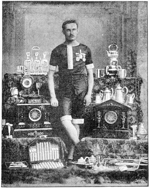 Schwarzes und weißes Foto eines Mannes in einem Fußballtrikot, der vor einem Tisch mit Pokale und Trophäen steht, die mit '1909-1910 WM-Sieger' beschriftet sind.