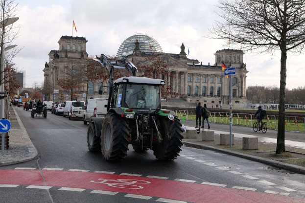 Ein grüner Traktor fährt am Reichstaggebäude in Berlin, Deutschland, vorbei, mit Fußgängern und Fahrradfahrern auf dem Gehweg, Bäumen, die die Straße säumen, und Flaggen auf dem Gebäude unter einem bewölkten Himmel.
