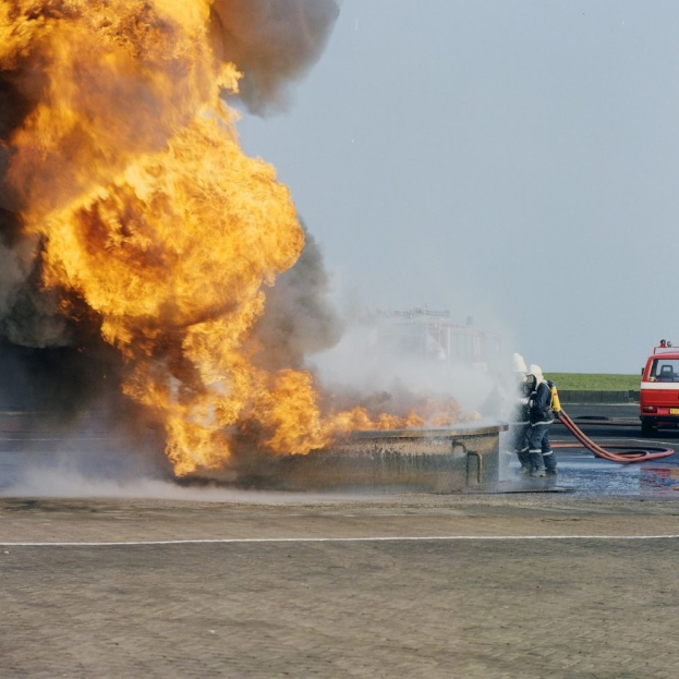 Feuerwehr auf der Straße von Flammen eingehüllt mit zwei Helmet tragenden Individuen, die Rohre halten, ein Fahrzeug im Hintergrund und der Himmel.