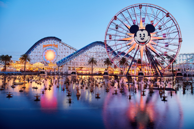 Eine lebendige Dämmerungsszene im Disneyland California Adventure mit einem riesigen Mickey-Maus-Rad im Vordergrund, einer Achterbahn im Hintergrund und Booten auf dem Wasser sowie einem Himmel aus Orange-, Pink- und Purpurfarben und silhouettenartigen Bäumen.