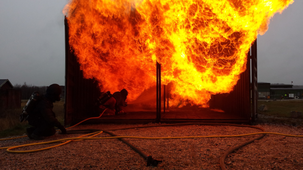 Feuerwehrleute löschen ein Gebäude Feuer mit einer auf dem Boden liegenden Leitung, umgeben von Schuppen, Bäumen und einem klaren blauen Himmel.