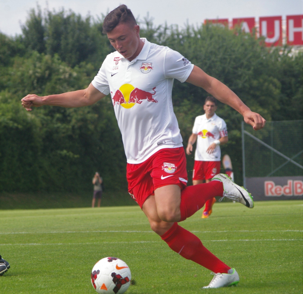 Ein professioneller Fussballspieler in einem roten und weissen Trikot kickt einen Fussball auf einem grasigen Feld mit Baumen und einem klaren blauen Himmel im Hintergrund, in der Nähe einer Tafel, auf der 'RB Leipzig' steht.