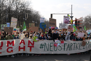 Eine große Gruppe von Menschen marschiert auf einer Straße, hält ein 'Menschliche Rechte'-Schild und verschiedene Plakate, wobei einige Masken tragen, unter einem klaren blauen Himmel mit Bäumen und Laternen im Hintergrund.