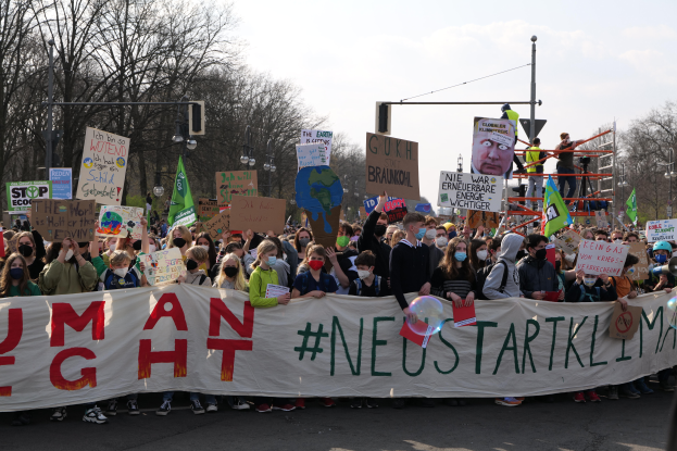 Eine große Gruppe von Menschen marschiert auf einer Straße, hält ein 'Menschliche Rechte'-Schild und verschiedene Plakate, wobei einige Masken tragen, unter einem klaren blauen Himmel mit Bäumen und Laternen im Hintergrund.