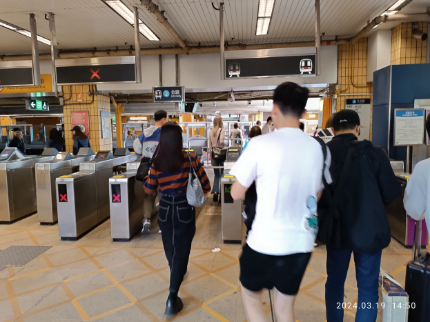 Menschen gehen durch einen Bahnhof mit Metalldetektoren, Hinweisschildern, Metallstangen und Deckenleuchten.