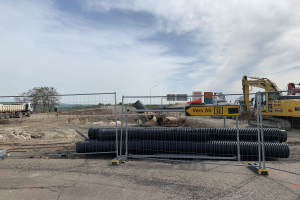 Eine Baustelle mit einem großen Haufen schwarzer Rohre in der Mitte, umgeben von einem Metallzaun, einem Bagger auf der rechten Seite, Fahrzeugen auf der linken Seite und Bäumen, Polen, Lampen und Schildern im Hintergrund.