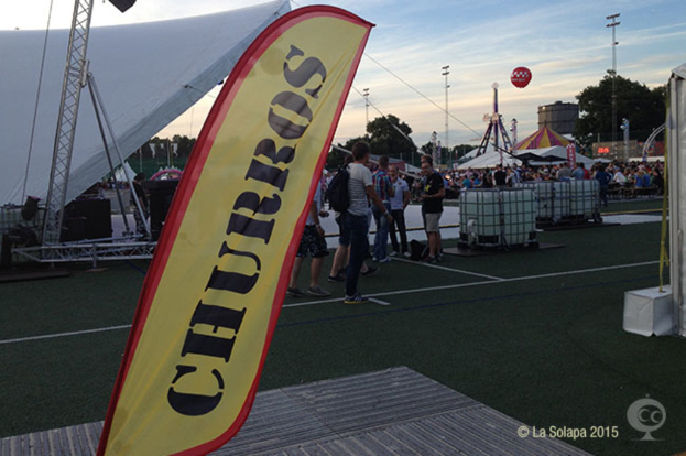 Eine KarnevalsSzene mit Zelten, einem zentralen Mast, Menschen, Bäumen, Strommasten, einem Himmel, Straßenbahnen auf der Straße und einem Eisenständer links.