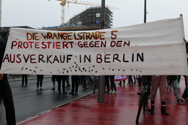 Eine Gruppe von Menschen steht auf einer Straße und hält ein Banner in der Hand, auf dem "Die Wrangellstraße 5 protestiert gegen den Ausverkauf in Berlin" steht. Im Hintergrund sind Fahrräder, Stangen, Gebäude und ein Kran zu sehen.