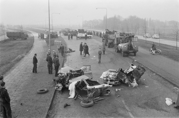 Schwarz-weiß-Szene eines Autounfalls am Straßenrand mit mehreren Fahrzeugen, darunter ein Landfahrzeug, und einer Gruppe von Menschen drumherum, mit Laternenpfählen, Bäumen und Himmel im Hintergrund.