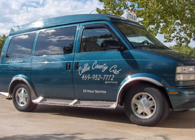 Blauer Van mit Text und Zahlen auf einem Parkplatz umgeben von Bäumen unter einem klaren Himmel, wahrscheinlich ein Taxi.