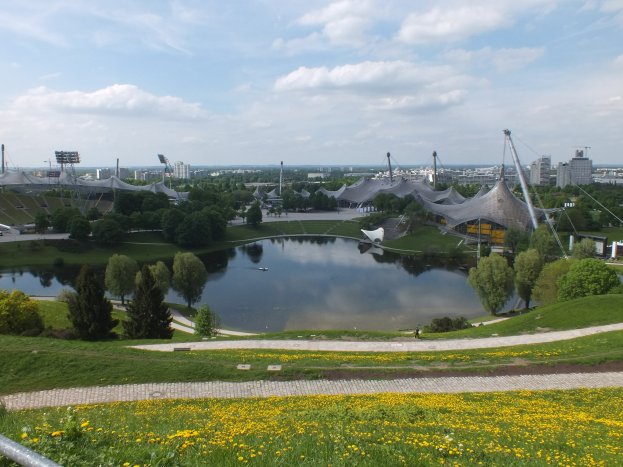 Ansicht des Olympiagel├Ąndes von einem Hügel aus, mit einem Teich im Vordergrund, umgeben von Gras, B├Ąumen und gelben Blüten, sowie Geb├Ąuden und Masten im Hintergrund unter einem klaren blauen Himmel.