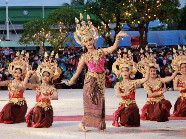 Eine Gruppe von Frauen in traditioneller Thai-Kleidung tanzt vor einem Publikum im Freien, mit Bäumen, Lichtern und einem Gebäude im Hintergrund bei klarem blauem Himmel.