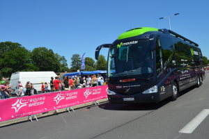 Ein schwarzer und grüner Bus fährt auf einer Straße neben einer Menschenmenge, einige tragen Mötzen, mit einer Fahne auf der linken Seite und Bäumen unter einem klaren blauen Himmel im Hintergrund.