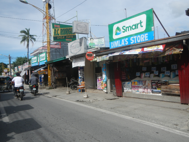 Eine belebte Straßenszene mit Menschen auf Motorrädern und Autos, umgeben von Gebäuden, Geschäften, Schildern, Strommasten, Kabeln, Straßenlaternen, Bäumen und einem bewölkten Himmel.