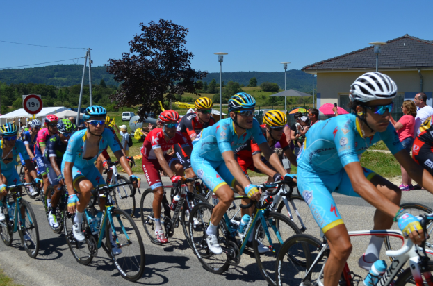 Gruppe von Radfahrern in Sportkleidung auf einer Straße mit Zelten, Fahrzeugen und Zuschauern im Hintergrund unter einem klaren blauen Himmel.
