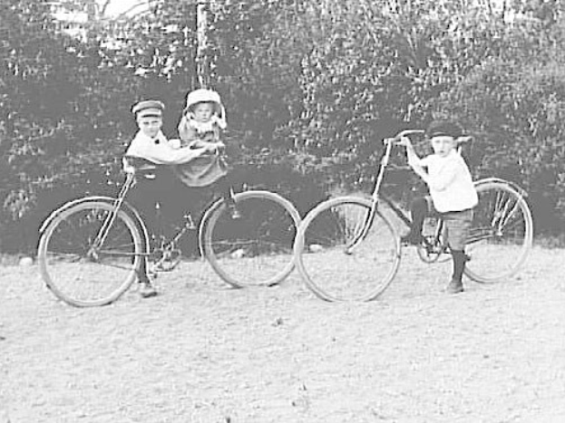 Zwei Kinder fahren Fahrräder in einem Park, eines hält ein Baby, mit Bäumen und einem Pfahl im Hintergrund.