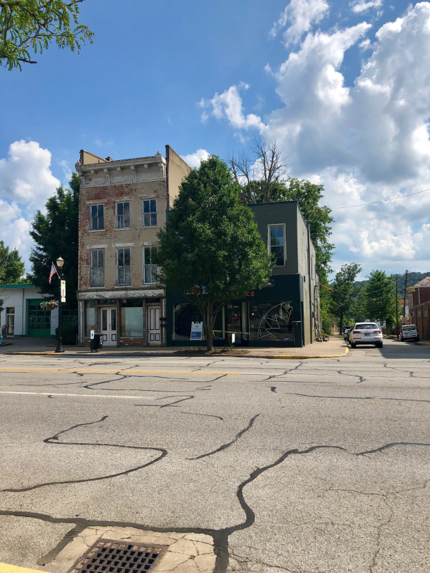 Eine Straße mit parkenden Autos, Gebäuden, Bäumen und Laternenmästen, mit einer beschädigten Straße und einem Gebäude im Vordergrund unter einem bewölkten Himmel.