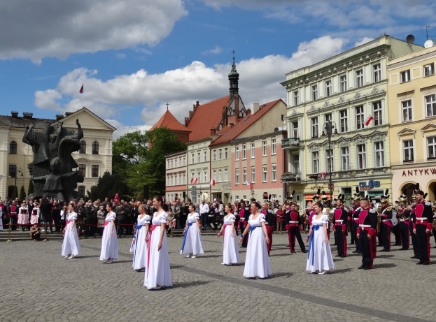 Eine Gruppe von Menschen in weißen Kleidern steht vor einer Menge mit Musikinstrumenten, mit Gebäuden, Bäumen, einer Statue, Flaggen und einem bewölkten Himmel im Hintergrund, wahrscheinlich während des Nationalfeiertags der Tschechischen Republik.